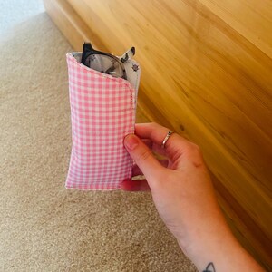 May include: A pink and white gingham eyeglasses case holding a pair of glasses. The case is made of fabric and has a soft, padded interior. The glasses have a black frame.