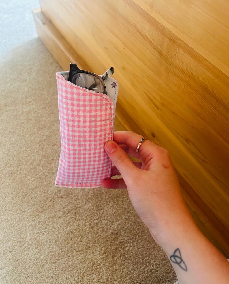 May include: A pink and white gingham eyeglasses case holding a pair of glasses. The case is made of fabric and has a soft, padded interior. The glasses have a black frame.