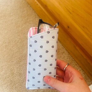 May include: A light blue eyeglasses case with a repeating navy anchor pattern. The interior lining is pink and white striped. Black framed eyeglasses are partially visible inside the case. The case is held by a hand.