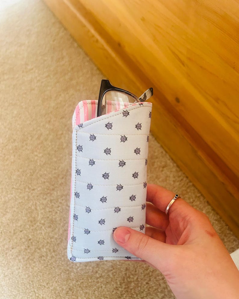 May include: A light blue eyeglasses case with a repeating navy anchor pattern. The interior lining is pink and white striped. Black framed eyeglasses are partially visible inside the case. The case is held by a hand.