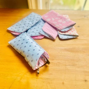 May include: A collection of fabric eyeglass cases in various patterns and colors. The cases are primarily blue and pink, with floral and gingham designs. One case holds a pair of eyeglasses. The cases are arranged on a wooden surface.