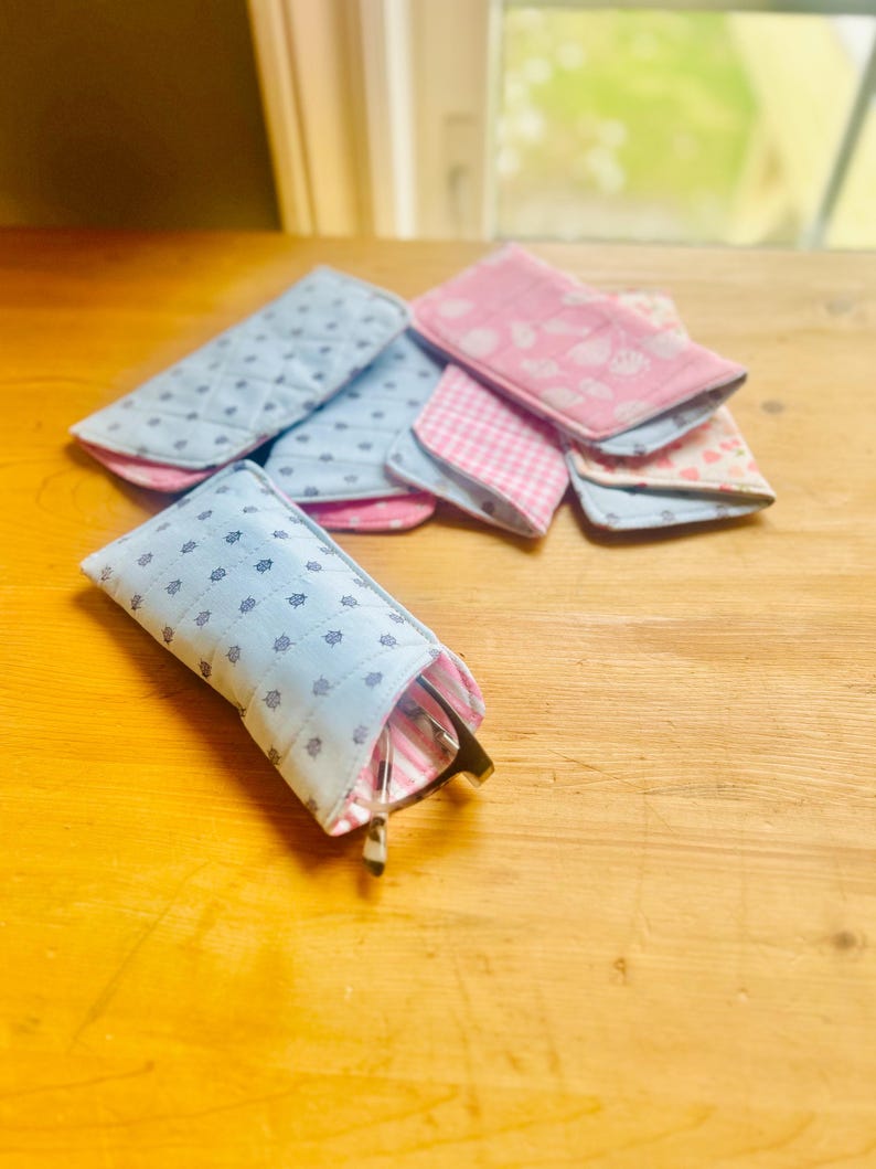 May include: A collection of fabric eyeglass cases in various patterns and colors. The cases are primarily blue and pink, with floral and gingham designs. One case holds a pair of eyeglasses. The cases are arranged on a wooden surface.