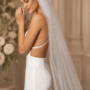 Puede incluir: Un velo de novia blanco largo hasta el suelo hecho de tela de tul transparente, adornado con peque&ntilde;as perlas blancas dispersas por toda la longitud. El velo est&aacute; sujeto al cabello y cae por la espalda, combinado con un vestido blanco sin mangas.