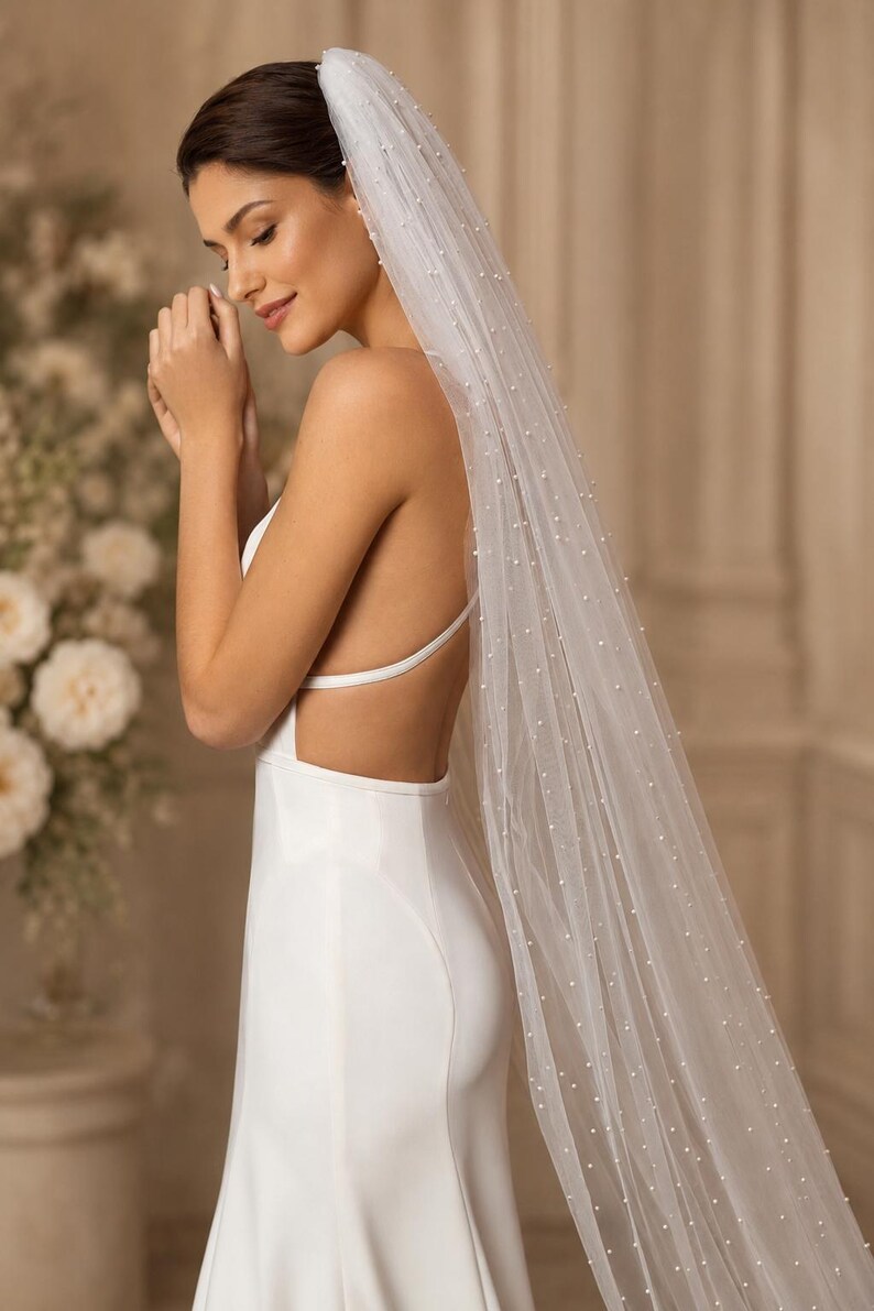 Puede incluir: Un velo de novia blanco largo hasta el suelo hecho de tela de tul transparente, adornado con peque&ntilde;as perlas blancas dispersas por toda la longitud. El velo est&aacute; sujeto al cabello y cae por la espalda, combinado con un vestido blanco sin mangas.