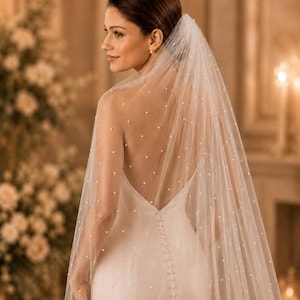 Puede incluir: Un velo de novia blanco transparente adornado con peque&ntilde;as perlas blancas dispersas, drapeado sobre los hombros y la espalda. El velo est&aacute; hecho de tela de tul fina y presenta una estructura delicada y ligera que cae por la espalda de un vestido.