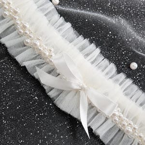 May include: A white garter featuring a ruffled tulle band adorned with a double row of white faux pearls and a central satin ribbon bow. The accessory is constructed from sheer, pleated fabric with a delicate texture and a consistent pearl trim.