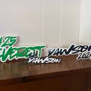 May include: Three custom-cut signs with the name "Everett Dawson" and the year "2024". The largest sign has the number "25" in green, with the name in green. The other signs are black and white.