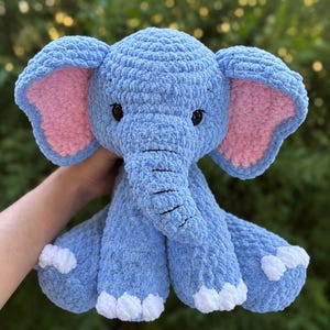May include: A handmade blue crochet elephant plush toy with pink inner ears and white accents on its feet. The elephant has black eyes and a trunk. The soft, cuddly toy is designed for children and is a charming addition to any nursery or playroom.