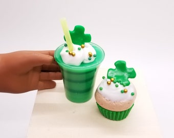 May include: A green beverage in a clear cup with a straw, topped with white foam, a green shamrock, and gold sprinkles. Beside it is a cupcake with white frosting, a green shamrock, and gold sprinkles. The items are on a white surface.