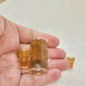 May include: Two miniature glass mugs filled with a light brown liquid, one is a larger mug with a handle, the other is a smaller mug. The mugs are held in a person's hand.