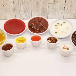 May include: A collection of eight small bowls filled with different types of soup. The bowls are white with a variety of soups, including red, yellow, brown, and white.