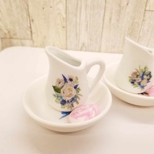 May include: Two white ceramic creamer pitchers with floral designs and a pink soap bar on a white saucer.