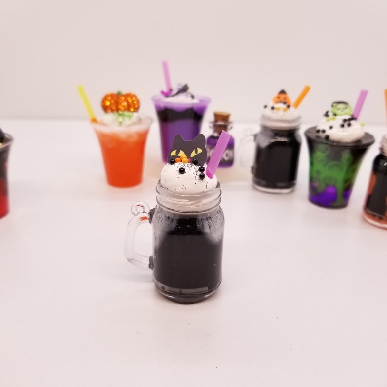 May include: A black and white Halloween themed mason jar with a black cat topper and a purple straw. The jar is filled with a black liquid and topped with white whipped cream. The jar is sitting on a white surface.