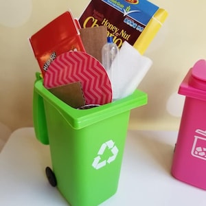 May include: A green toy recycling bin with a white recycling symbol on the side. The bin is filled with various items, including a red box with the word "MEGASIZE" on it, a brown cardboard box, a red and white patterned circle, a white paper towel, a clear plastic bottle, and a yellow box with the words "Honey Nut Cheerios" on it.