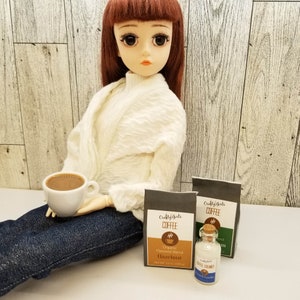 May include: A doll holding a small white cup of coffee. Two small bags of coffee are on the table, one labeled "Crafty Girls Coffee Hazelnut" and the other labeled "Crafty Girls Coffee". A small bottle of coffee creamer is in front of the bags.