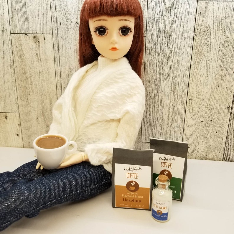 May include: A doll holding a small white cup of coffee. Two small bags of coffee are on the table, one labeled "Crafty Girls Coffee Hazelnut" and the other labeled "Crafty Girls Coffee". A small bottle of coffee creamer is in front of the bags.