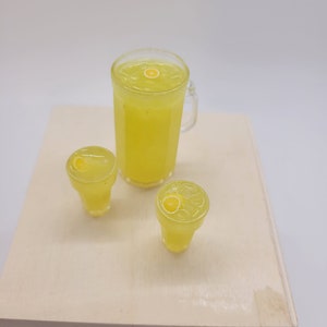 May include: Three glass containers filled with yellow lemonade. The largest container is a pitcher with a handle. The two smaller containers are glasses. Each container has a lemon slice floating in the liquid.
