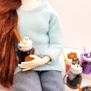 May include: A doll holding a mason jar filled with a chocolate drink topped with whipped cream and a ghost-shaped marshmallow. There are other mason jars filled with colourful drinks and Halloween-themed decorations.