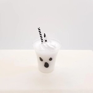 May include: A white ghost-shaped milkshake with black eyes and a mouth in a clear plastic cup with a black and white striped straw.
