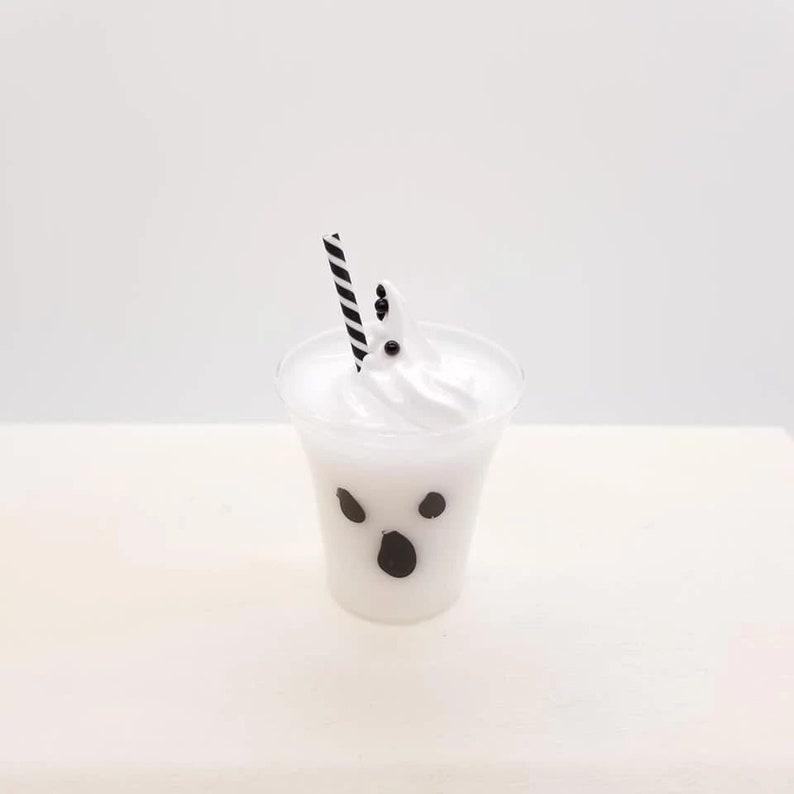 May include: A white ghost-shaped milkshake with black eyes and a mouth in a clear plastic cup with a black and white striped straw.