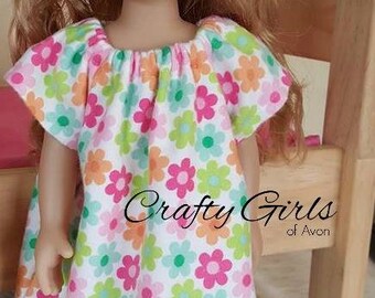 May include: A doll wearing a floral print dress with pink lace trim. The dress features a pattern of pink, orange, and green flowers on a white background. The doll has blonde hair and blue eyes. Text on the dress reads "Crafty Girls of Avon."
