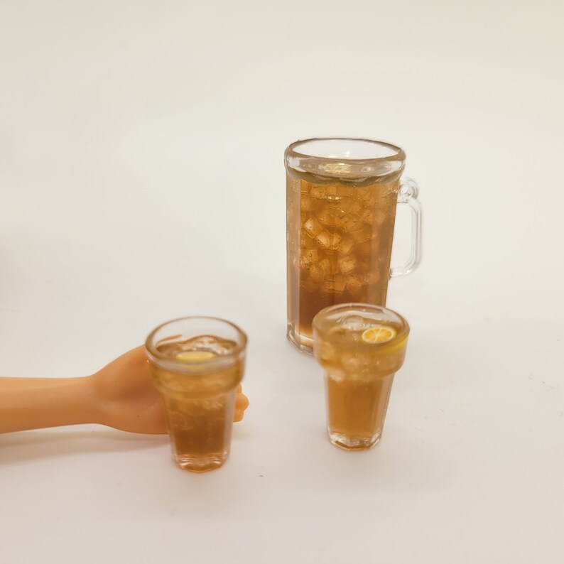 May include: Three clear glass cups filled with iced tea. The largest cup has a handle and is filled with ice and brown liquid. The two smaller cups are filled with ice and brown liquid and have a slice of lemon in each.