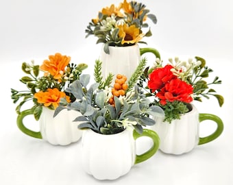 May include: Four white ceramic pumpkin-shaped mugs with green handles, each containing a different artificial floral arrangement. The arrangements feature orange, yellow, and red flowers, green foliage, and small orange berries in a cluster.