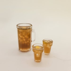 May include: A glass pitcher filled with iced tea and two glasses of iced tea with lemon slices. The pitcher and glasses are clear and the tea is a light brown color.