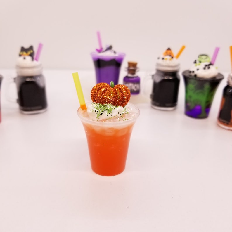 May include: A miniature orange pumpkin sits atop a white-topped orange drink with a green straw in a clear plastic cup. The drink is surrounded by other miniature Halloween-themed drinks in various colours and designs.