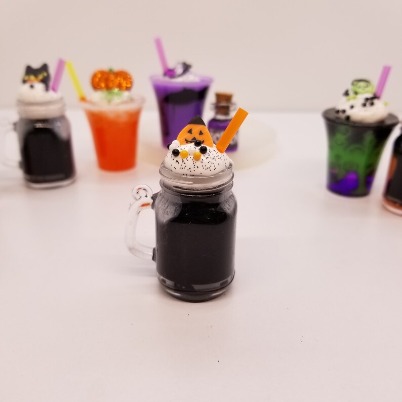 May include: A black and white Halloween-themed milkshake in a mason jar with a pumpkin-shaped candy topping and an orange straw. The milkshake is surrounded by other Halloween-themed drinks.