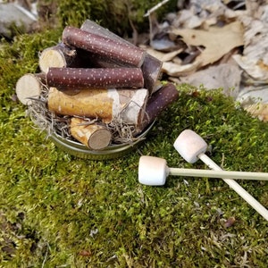 May include: A miniature campfire scene with a metal container filled with small logs of varying colors and textures. Two toasted marshmallows on skewers rest on a bed of green moss, creating a cozy, playful aesthetic.