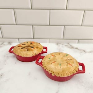 May include: Two miniature pot pies in red ceramic dishes. The pies have a golden-brown crust with decorative slits and visible vegetable pieces. Each dish has two handles. The background is a white tile wall and a marble surface.