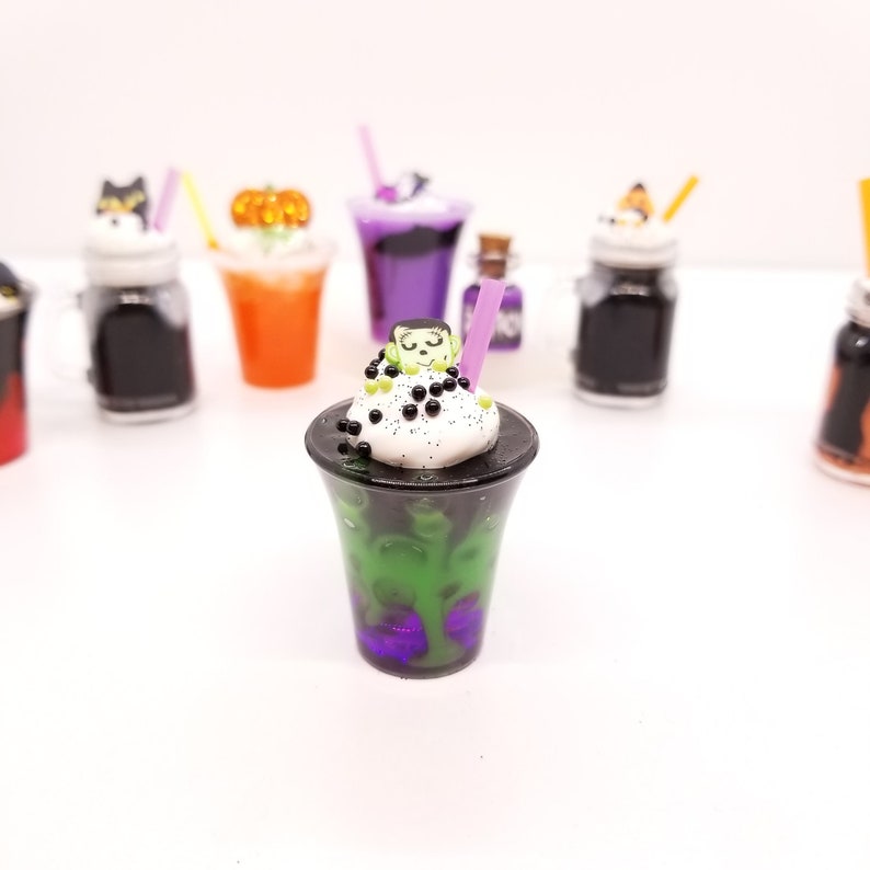 May include: A green, black and white Halloween-themed milkshake with a purple straw and a small Frankenstein face on top. The milkshake is in a clear plastic cup with a purple rim.
