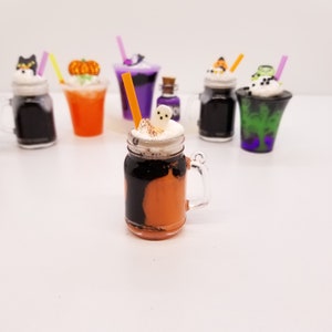 May include: A Halloween-themed orange and black milkshake in a mason jar with a ghost marshmallow on top. The milkshake is topped with whipped cream and a sprinkle of cinnamon. There are other Halloween-themed milkshakes in the background.