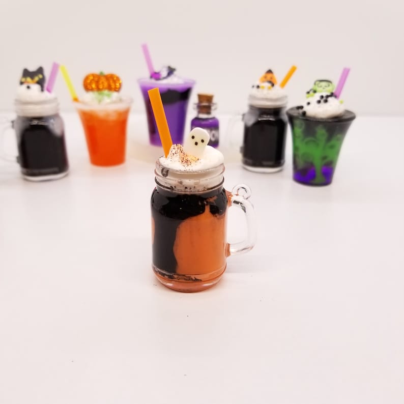 May include: A Halloween-themed orange and black milkshake in a mason jar with a ghost marshmallow on top. The milkshake is topped with whipped cream and a sprinkle of cinnamon. There are other Halloween-themed milkshakes in the background.