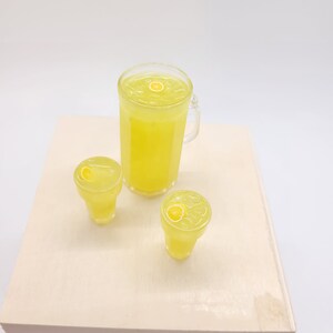 May include: A set of three clear glass containers filled with yellow lemonade. The containers are a pitcher and two glasses. Each container has a lemon slice floating on top.