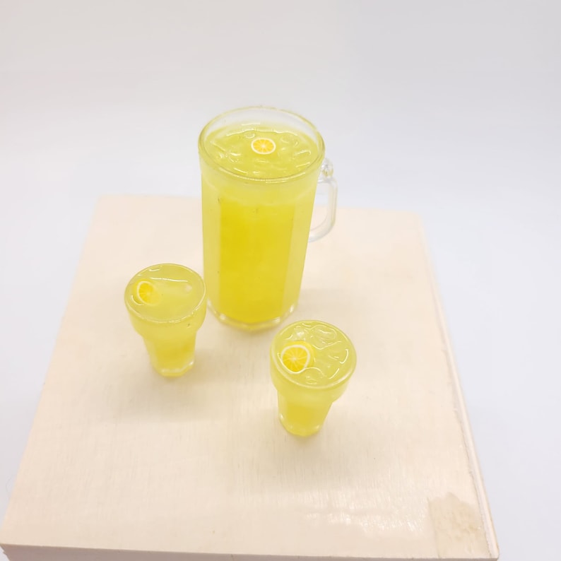 May include: A set of three clear glass containers filled with yellow lemonade. The containers are a pitcher and two glasses. Each container has a lemon slice floating on top.