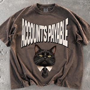Op de afbeelding: Een donkerbruin t-shirt met korte mouwen en een afbeelding van een zwarte kat die een wit overhemd met kraag en een zwarte stropdas draagt. Boven de kat staat de tekst ACCOUNTS PAYABLE in grote, dikke, witte hoofdletters gedrukt.
