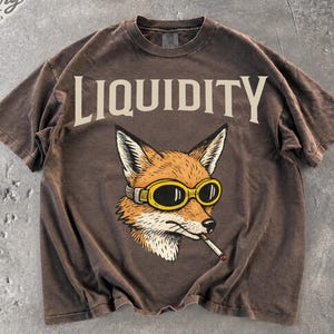 Può includere: Una t-shirt a maniche corte marrone scuro con la parola LIQUIDITY in grandi lettere color crema sul petto. Sotto il testo si trova un'illustrazione grafica di una volpe che indossa occhiali con montatura gialla e tiene una sigaretta in bocca.