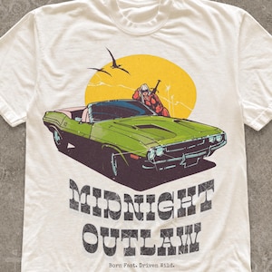 May include: An off-white short-sleeve t-shirt featuring a graphic of a green muscle car, a person, and a yellow sun. The text reads "MIDNIGHT OUTLAW" and "Born Fast. Driven Wild." The design has a distressed, vintage-inspired texture throughout.