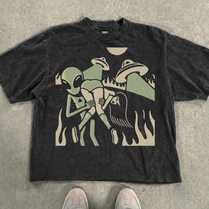 Könnte beinhalten: Ein schwarzes Kurzarm-T-Shirt mit einem Grafikdruck, der eine grüne Alien-Figur zeigt, die eine Person trägt. Das Design umfasst zwei fliegende Untertassen, einen Halbmond und flammenartige Formen in gedeckten Grün- und Cremetönen auf der Brust.