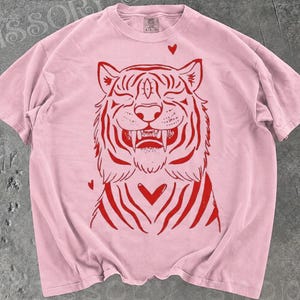 May include: A light pink short-sleeve t-shirt featuring a red graphic print of a tiger face with a third eye on its forehead, bared fangs, and small heart shapes surrounding the tiger. The shirt has a crew neckline and a standard fit.