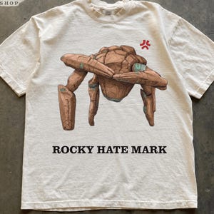 May include: An ivory short-sleeve t-shirt featuring a brown rock creature illustration with teal accents, a red anger symbol, and the text ROCKY HATE MARK printed in black. The shirt has a crew neckline and a relaxed fit.