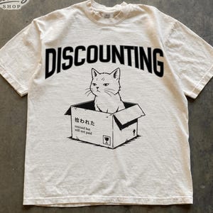 May include: An ivory short-sleeve t-shirt featuring a black graphic of a cat sitting inside a cardboard box. The word DISCOUNTING is printed in a large black arch above the cat. The box includes the text 拾われた, rescued but still not paid.