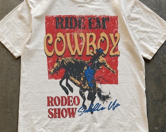 May include: An ivory short-sleeve t-shirt featuring a distressed red rectangular graphic with the text RIDE EM COWBOY and RODEO SHOW. The graphic depicts a cowboy riding a bucking horse in black, tan, and blue colours with the script text Saddle Up.
