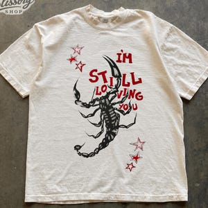 May include: An ivory short-sleeve t-shirt featuring a black graphic of a scorpion in the centre. The red text reads "I'M STILL LOVING YOU" integrated with the scorpion. Six red outlined stars are positioned around the graphic design.