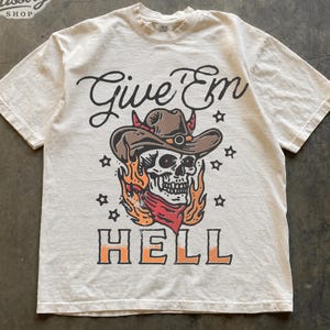 May include: An ivory short-sleeve t-shirt featuring a graphic print of a skull wearing a brown cowboy hat and a red bandana, surrounded by orange flames and black stars. The text "Give 'Em" appears above the skull, and "HELL" is printed below.