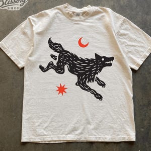 May include: An ivory short-sleeve t-shirt featuring a black graphic of a running wolf, a red crescent moon, and a red eight-pointed star. The text Blissory SHOP is printed in the top left corner. The shirt has a crew neckline and a relaxed fit.