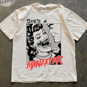 May include: An ivory short-sleeve t-shirt featuring a black and white comic-style graphic of a person with a monstrous, toothy mouth. The text reads "She's a" in black and "MANEATER" in distressed red lettering below the illustration.