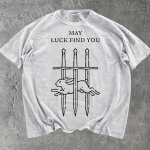 May include: A light grey t-shirt featuring the text "MAY LUCK FIND YOU" above a graphic of a rabbit running through a sword-barred gate. The design is in black ink.